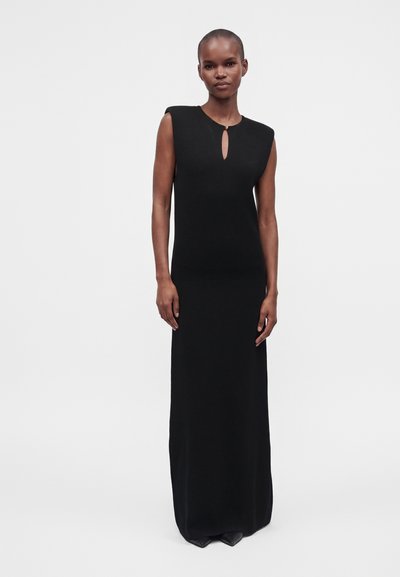 Femme debout portant une robe noire longue sans manches avec des épaulettes et un petit décolleté en forme d'œillet fermé par un seul bouton.
