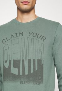 Sudadera verde con cuello redondo que presenta una gran impresión gráfica que dice "RECLAMA TU MEZCLILLA" en negro, con un aspecto texturizado.