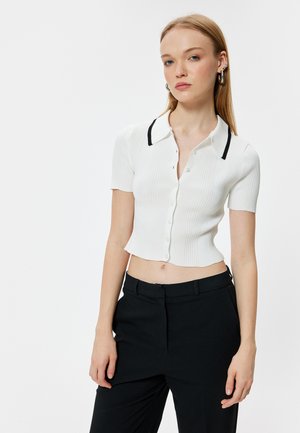 Top blanco de manga corta con relieve y cuello ribeteado en negro, que presenta un cierre delantero con botones y un dobladillo recortado, combinado con pantalones negros.