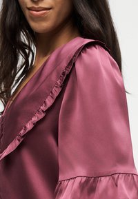 Femme portant un chemisier mauve brillant avec un bord volanté au col et des manches longues bouffantes, visible du cou au milieu du torse.