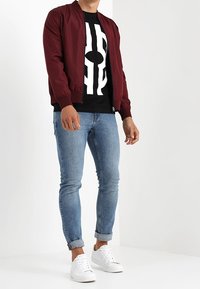 Chaqueta bomber burdeos sobre una camiseta gráfica negra, combinada con jeans ajustados azules y zapatillas blancas. Diseño sencillo con elementos informales.