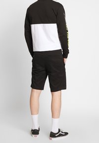 Zwart shirt met lange mouwen, voorzien van een wit paneel en gele tekst, gecombineerd met zwarte short. Model draagt witte sokken en zwarte sneakers.