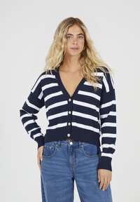 Vrouw met lang blond haar, gekleed in een marineblauwe en witte gestreepte knooptrui en blauwe jeans, staande met één hand op de heup.