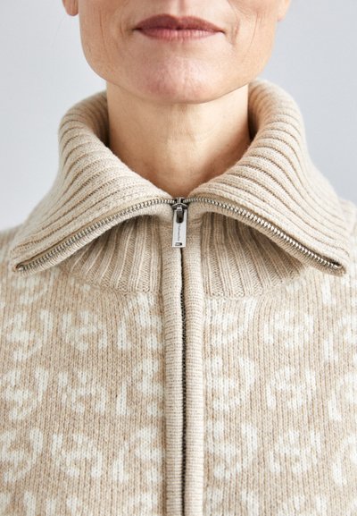 Beige stickad jacka med räfflad krage och dragkedjestängning, med ett texturerat mönster i ljusare beige. Mjuk, varm tyg med en mysig design.