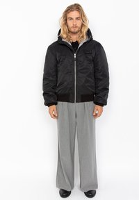 Zwarte gewatteerde jas met ritssluiting, elastische manchetten en een gevoerde capuchon, gedragen over grijs gestreepte broek en zwarte schoenen.