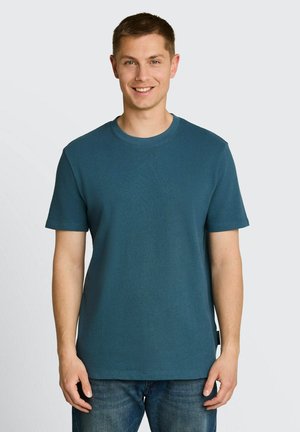 Junger Mann lächelnd, trägt ein einfarbiges blaugrünes kurzärmeliges Hemd und blaue Jeans, steht vor einem hellgrauen Hintergrund.