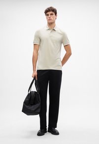 Polo beige clair avec un petit logo, associé à un pantalon noir à jambes droites et un sac de sport en tissu noir, sur un fond neutre.