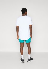Witte korte mouwen shirt met een afgeronde zoom, gecombineerd met turquoise shorts. De persoon draagt zwarte sportschoenen en witte sokken.