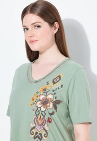Groene T-shirt met korte mouwen en een V-hals, voorzien van kleurrijke bloemenborduurwerk en decoratieve stiksel langs de halslijn.