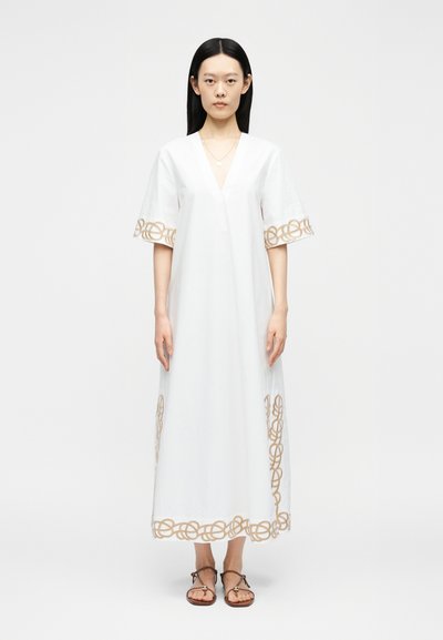 Femme aux cheveux noirs et raides portant une robe blanche longue jusqu'aux chevilles avec une bordure à motif chaîne beige et des sandales marron, debout sur un fond blanc.