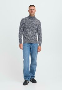 Moški stoji oblečen v sivo melirano pulover s turtleneck ovratnikom, svetlo modre jeans hlače in črne teniske z belimi črtami, na enobarvni beli podlagi.