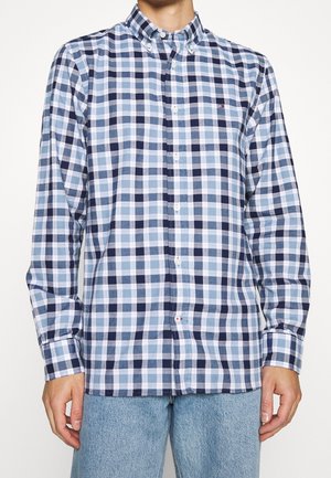 Mężczyzna ubrany w koszulę na guziki z długim rękawem w niebiesko-białą kratę oraz jasnoniebieskie dżinsy, stojący na jednolitym tle.