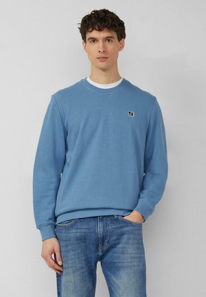 Giovane uomo con capelli scuri e ricci che indossa una felpa blu sopra una camicia bianca e jeans blu, in piedi davanti a uno sfondo grigio uniforme.