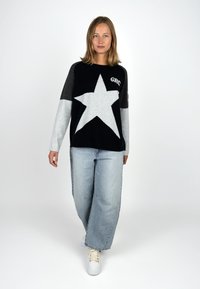 Black and gray long-sleeve sweater with a white star, featuring "GRC" embroidery. Paired with light blue wide-leg jeans and white sneakers.