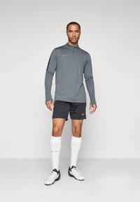 Sivi dugih rukava sportski majica s ružičastim Nike logom, uparena s crnim šortsem s narančastim Nike logom. Bijele čarape i kopačke upotpunjuju odjevnu kombinaciju.