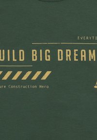 Dunkelgrünes T-Shirt mit gelbem Text, auf dem steht "GROßE TRÄUME BAUEN" und einem baustellenbezogenen grafischen Motiv mit Warnstreifen. Weiche Stoffstruktur.