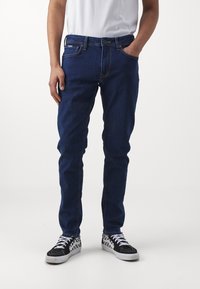 Dark blue denim jeans with a slim fit, five pockets, and contrast stitching. Worn with black and white checkered sneakers.
