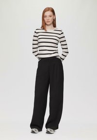 Haut à manches longues rayé en crème et noir, associé à un pantalon noir ample. Le modèle porte des chaussures de sport grises. Design simple et moderne.