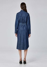 Femme portant une robe midi en denim bleu foncé à manches longues avec une ceinture, tournée de dos, accompagnée de sandales à talons hauts noires.
