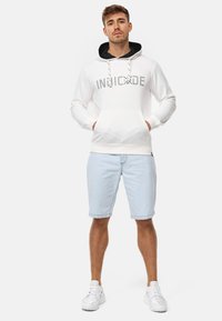 Haut à capuche blanc avec bord noir et texte graphique argenté, associé à un short bleu clair et des baskets blanches. Il dispose d'une poche kangourou et de cordons.