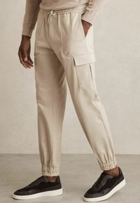 Pantalons cargo beiges avec poignets et taille élastiques, comportant des poches latérales et un tissu texturé. Associés à des baskets noires à enfiler.