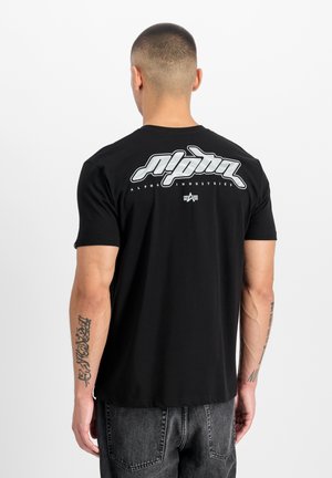Mann mit kurzem Haar trägt ein schwarzes Alpha Industries T-Shirt mit weißem Logo auf dem oberen Rücken, kombiniert mit dunkelgrauen Jeans, steht mit dem Rücken zur Kamera.