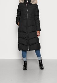 Abrigo puffer largo negro con capucha forrada de piel sintética, con detalles de acolchado horizontal, cremallera frontal completa y bolsillos laterales.