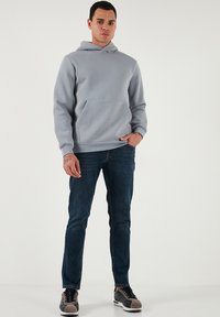 Szürke kapucnis pulóver, elülső zsebbel, kerek zsinóros kapucnival, bordás mandzsettákkal, sötétkék farmerral és szürke sportcipővel párosítva. Sima szövettextúra.