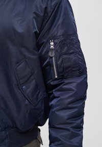 Sleevy námořnické modré bomberové bundy s kapsou na zip a kapsou na knoflík, nosí se přes olivově zelené kalhoty na šedém pozadí.