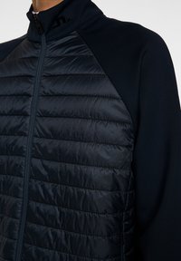 Närbild av en mörkblå jacka med vadderad front, hög krage och dragkedjestängning, som framhäver dess texturerade tyg och sportiga design.