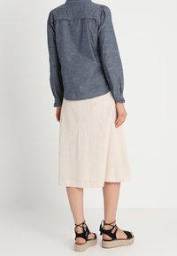 Chemise légère bleu foncé à manches longues avec un dos plissé, associée à une jupe beige en A et des espadrilles noires à lacets.