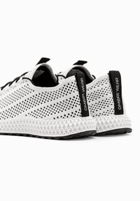 Paire de chaussures de sport blanches en maille avec des languettes noires portant l'inscription "OMBRE WEAR" et des semelles blanches texturées sur fond blanc.