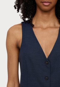 Marinblå ärmlös klänning med djup V-halsringning, med två svarta knappar framtill och en texturerad tygfinish.