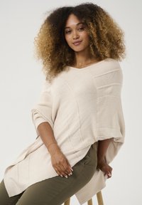 Helles beiges gestricktes Poncho mit diagonalen Mustern, weiten Ärmeln und einer lässigen Passform, kombiniert mit olivgrünen Hosen. Akzentdetails aus goldener Schmuck.
