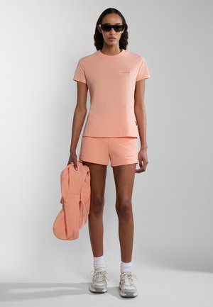 Pfirsichfarbene Baumwoll-T-Shirt- und Shorts-Set, mit einem kleinen Logo auf der Brust und an der Seite. Modell hält eine passende Jacke. Sonnenbrille getragen.