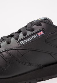 Baskets noires Reebok avec un design à lacets, des coutures détaillées et le drapeau Union Jack à côté du logo Reebok sur le côté.