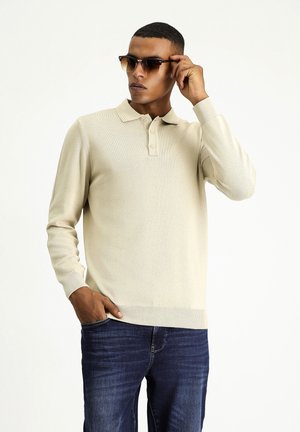 Pull en polo beige à manches longues avec un plastron à trois boutons, porté avec un jean bleu foncé. La texture semble lisse avec une finition douce.