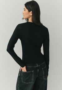 Schwarzes Langarmshirt aus weichem Stoff, figurbetontes Design, kombiniert mit dunklen Jeans mit großen Gesäßtaschen und einem verblassten Muster.
