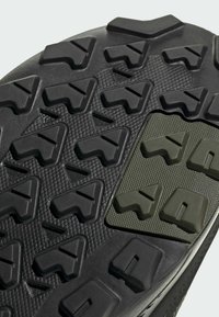 Schwarze Gummisohle mit strukturiertem Grip-Muster und angularen Stollen. Enthält einen grünen strukturierten Akzentbereich mit ähnlichen Designelementen.