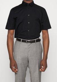 Černá košile s krátkými rukávy na knoflíky s límečkem, zdobená lesklými černými knoflíky, nošená s světle šedými formálními kalhotami a černým opaskem.