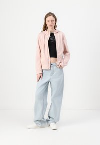 Ανοιχτό ροζ μπουφάν με φερμουάρ και κουκούλα, συνδυασμένο με ένα μαύρο κοντό τοπ και ανοιχτό μπλε παντελόνι σε φαρδιά γραμμή. Λευκά sneakers ολοκληρώνουν την εμφάνιση.