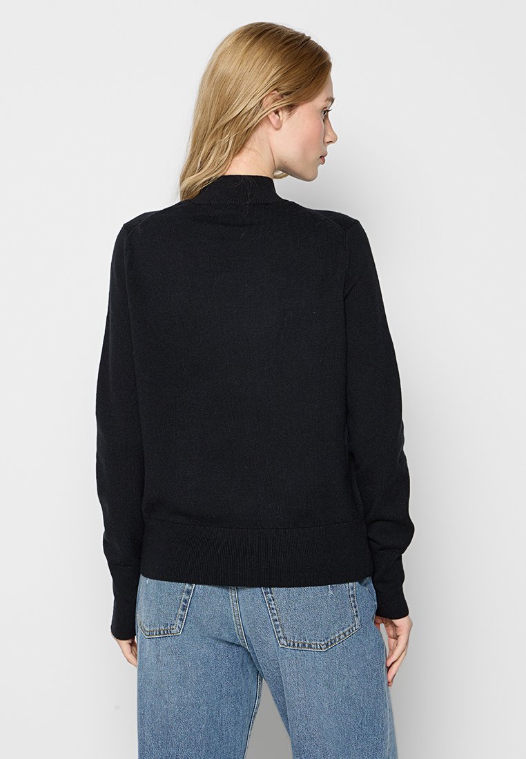 Schwarzer Strickpullover mit rundem Ausschnitt und gerippten Bündchen. Der Rücken hat eine glatte Textur und eine taillierte Passform. Kombiniert mit hellblauen Jeans.