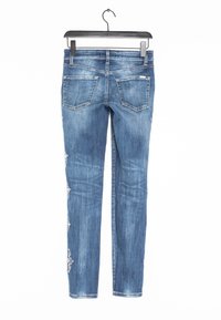 Blå denimjeans hängande från en svart galge. Har en smal passform, ljus slitage och vita broderade mönster på sidorna.