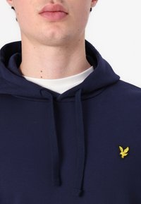 Felpa a collo alto di colore navy con coulisse, realizzata in tessuto morbido. Presenta un piccolo logo ricamato giallo sul petto. Texture liscia.