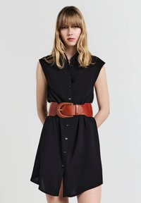 Giovane donna con capelli biondi che indossa un vestito nero senza maniche con bottoni e una larga cintura di pelle marrone su uno sfondo semplice.
