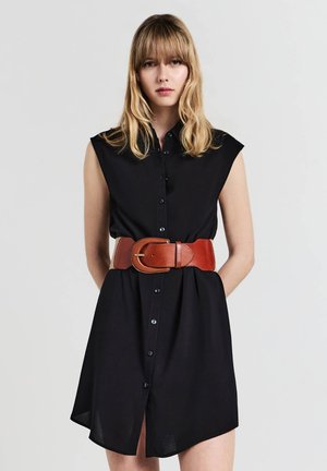 Giovane donna con capelli biondi che indossa un vestito nero senza maniche con bottoni e una larga cintura di pelle marrone su uno sfondo semplice.