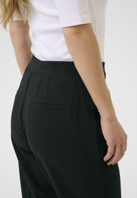 Schwarze Hose mit strukturiertem Finish, ausgestattet mit einem elastischen Bund und einer einzelnen Gesäßtasche. Das Outfit wird durch ein weißes, figurbetontes Oberteil ergänzt.