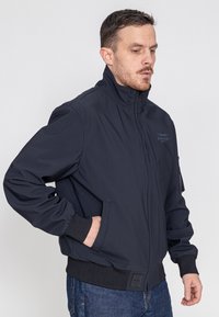 Marineblaue Bomberjacke mit hohem Kragen, Reißverschluss, gerippten Bündchen und zwei Seitentaschen. Hergestellt aus glattem, robustem Material mit minimaler Branding.
