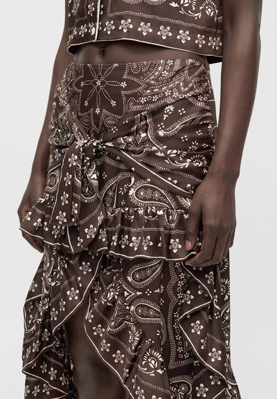 Personne à la peau foncée portant une jupe marron à motif cachemire avec des volants et un nœud noué à la taille, associée à un top court assorti.