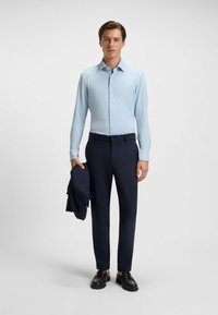 Helles blaues Langarmhemd mit durchgehender Knopfleiste, kombiniert mit dunkelblauen Hosen. Als Accessoires wird ein marineblauer Blazer in einer Hand gehalten.